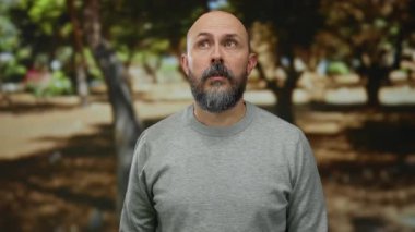 Şaşırmış bir ifadeyle parkta dikilen adam, gri bir süveter giyiyor, ağaçların gölgelerinin altında açık havada, ilginç bir an sergiliyor..