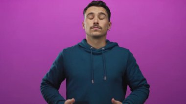 Mavi kapüşonlu rahat yüzü olan, gözleri kapalı genç İspanyol bir adam canlı pembe arka plan üzerinde sanki meditasyon yapıyor ya da tecritte sakinleşiyormuş gibi el kol hareketi yapıyor.