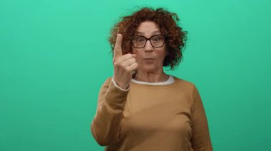 Kıvırcık saçları ve gözlükleriyle duygularını gösteren, kahverengi bir gömlek giyen ve çerçevede izole edilmiş bir kadın, canlı yeşil bir arka plana karşı ifade edici bir poziyonda parmaklarıyla jestler yapıyor..