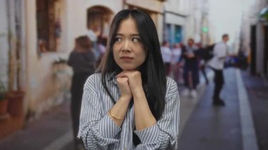 Genç bir kadın, hareketli bir sokakta, parlak bir günde çizgili gömleğiyle dalgın dalgın duruyor..