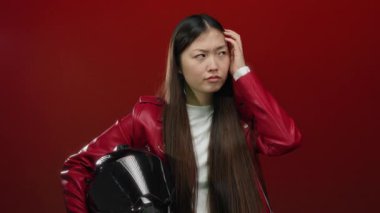 Kırmızı ceketli bir kadın kırmızı arka planda miğfer tutuyor düşünceli ve odaklanmış görünüyor.