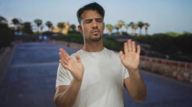Güneşli bir günde, arka planda palmiye ağaçları olan, kararsızlığını ve ince bir kafa karışıklığını ifade eden Latin kökenli genç açık havada belirsiz bir hareket sergiliyor..