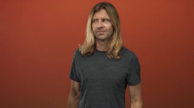 Sarışın adam kaşlarını çatar ve turuncu stüdyoda burnunu çimdikler. Gri gömlekli, uzun saçlı, omuzlarının üzerinde tiksinti vardır..