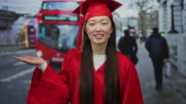 Kırmızı mezuniyet elbisesi giymiş, arka planda insanlar ve otobüslerle gülümseyen bir kadın, bir başarı ve kültürel çeşitlilik hissi sergiliyor..