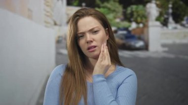 Güneşli bir şehir sokağında, taş bir binanın yanında, mavi kazak giymiş, yanaklı bir kadın. Acı..