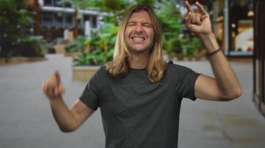 Uzun sarı saçlı, sırıtan bir adam sokakta taş boynuzlarını göstermek için iki kolunu kaldırıyor..