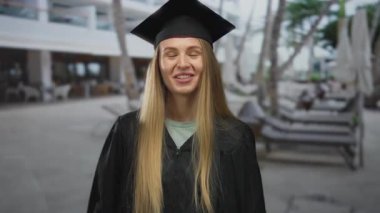 Şehir dışında mezuniyeti kutlayan kadın, kep ve cübbe giymiş, parlak bir günde arka planda yeşillikle neşeyle gülümseyen kadın..