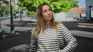 Çizgili tişörtlü genç sarışın kadın güneşli bir sokakta poz veriyor. Olumlu ve kendinden emin bir tavır sergiliyor. Arka planda şehir manzarası var..