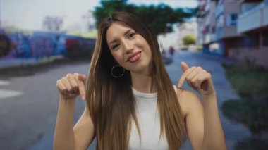 Beyaz atletli genç İspanyol kadın güneşli şehir caddesinde parmaklarını aşağı doğru gösteriyor; dostça bir davet..
