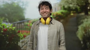Yeşil bir parkta boynunda sarı kulaklıklarla gülümseyen İspanyol adam, kollarını yana doğru uzatıyor, rahatlamayı ve dışarıda eğlenmeyi yansıtıyor..