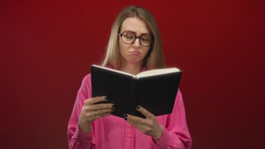 Gözlüklü ve pembe gömlekli bir kadın canlı kırmızı bir arka plana karşı kitap tutuyor, düşünceli ve düşünceli görünüyor..