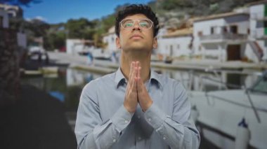 Man with clasped hands, closed eyes, and glasses at port next to white boat under clear sky and bright sunlight; hope.