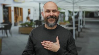 Sakallı bir adam şehir merkezindeki bir restoranın terasında jest yapıyor..