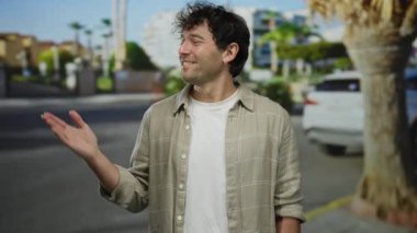 İspanyol bir adam sokakta gülümsüyor, avucunu uzatıyor, kentsel arka planı ve günlük kıyafetleriyle, güneşli bir günde hoş geldin jesti sergiliyor..