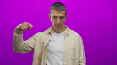 Tarafsız ifadeli bir adam parlak pembe arka plana işaret ediyor. Günlük bej gömlek giyiyor..