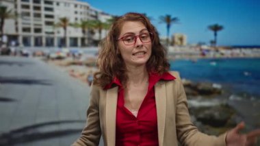 Kırmızı gözlüklü ve bej elbiseli bir kadın deniz kenarında mavi gökyüzü ve palmiye ağaçlarıyla geziniyor plaj ortamında karışık duygular gösteriyor..