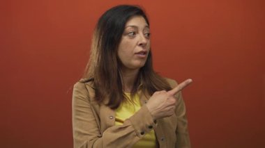 Orta yaşlı bir kadın, el hareketlerini vurgulayan turuncu bir stüdyo odasında yan yan bakarken parmağıyla objeyi işaret ediyor..