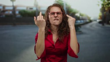 Kalabalık bir caddede kırmızı gömlek ve gözlük takan, şehir ortamında elleriyle ve yüz ifadesiyle duygularını gösteren bir kadın..