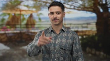 Tişörtlü genç adam kameraya emin adımlarla işaret ediyor. Arka planda bulanık manzaralı bir açık hava parkının ortasında duruyor.