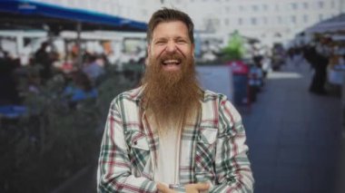 Uzun sakallı genç adam gözleri kapalı güler ve açık hava restoranının terasında kareli gömlek giyerek karnını tutar; neşe kahkahası.