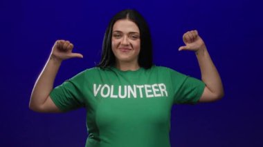 Kendine güvenen, yeşil tişörtlü, gülümseyen ve kendine mavi arka planda bakan gönüllü genç bir kadın.