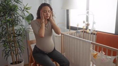Hamile bir kadın, bebek eşyaları, beşik ve bitkilerle çevrili bir bebek odasında rahatsızlık yaşıyor. Hamile bir annenin günlük hayatındaki samimi bir anı gösteriyor..