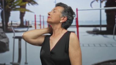 Gri saçlı, kolsuz bir elbise giyen, ensesinde bulanık plaj ve fitness ekipmanları olan bir deniz kenarında düşünceli bir şekilde dikilirken boynuna dokunan yaşlı bir kadın..