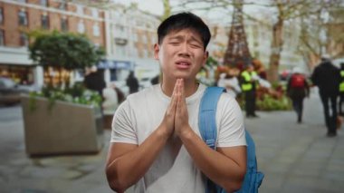 Sırt çantalı genç adam, kalabalık bir şehir caddesinde elleri sımsıkı bağlanmış bir şekilde, sanki şehir dışında dua ediyormuş gibi duruyor..