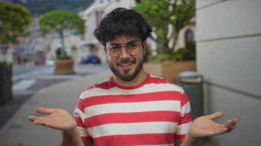 Sakallı genç adam canlı bir sokak ortamında gülümsüyor, gözlük takıyor ve kırmızı çizgili bir gömlek giyiyor, mutluluk ve rahatlama taşıyor..