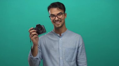 Gözlüklü genç adam kamerayı tutuyor ve yeşil arka planda barış işareti yapıyor. Sıradan bir tarz ve mutluluk sergiliyor..