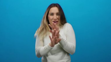Beyaz tişörtlü genç sarışın kadın, izole bir ortamda parlak mavi bir duvara şakacı hareketlerle el hareketleriyle ve doğal bir ifadeyle duygularını ifade ediyor..