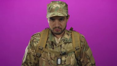 Yeşil kamuflaj üniforması, sırt çantası askısı ve künye giyen asker mor stüdyoda gözlerini dikiyor..