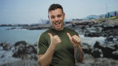 Dövmeli ve yeşil gömlekli genç adam gülümsüyor ve arka planda bulutlu bir gökyüzü ve sakin bir okyanus olan kayalık bir sahil bölgesinde baş parmağıyla geriye bakıyor..