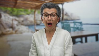 Gri saçlı yaşlı kadın deniz kıyısında, arka planda deniz ve kumsal gezintisine şaşırmış görünüyor, rahatlatıcı bir yürüyüş sırasında merak ve keyif karışımı taşıyor..