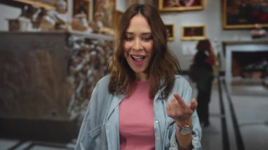Tablolar ve heykellerle çevrili bir sanat galerisinin içinde gülümseyen İspanyol kadın pembe gömlek ve kot ceket giyerken el hareketleriyle şaşkınlığını dile getiriyor..