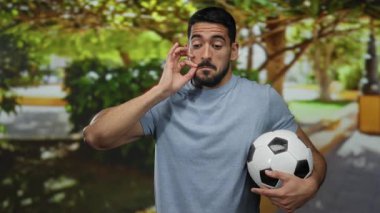 Futbol topu tutan sakallı genç bir adam güneşli bir açık hava parkında düşünceli bir ifade sergiliyor..