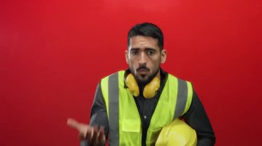 Kırmızı arka planda kaskı ve kulaklığı olan, güvenlik yeleği giymiş genç İspanyol bir adam, portre çerçevesinde endişeli ve anlamlı görünüyor.