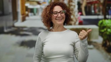 Orta yaşlı İspanyol kadın, şehir sokaklarında sırıtan gözlüklü, kıvırcık esmer saçlı, sıradan giyinen ve pozitifliğini baş parmaklarıyla ifade eden bir kadın..