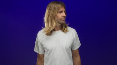 Sarışın ve uzun saçlı bir adam kobalt mavisi stüdyo ortamında sağa doğru işaret ediyor..