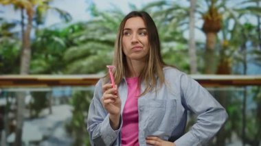 Parkta elinde pembe jiletle dolaşan, palmiye ağaçları olan, pembe gömlekli, mavi ceketli genç bir kadın.