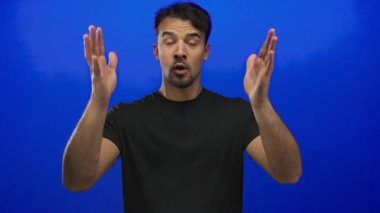 Genç İspanyol asıllı bir adam kameraya karşı konuşurken öfkesini ifade ediyor. Mavi arka planda, duyguları vurguluyor ve video içeriği oluşturulması için izole bir ortam yaratıyor..