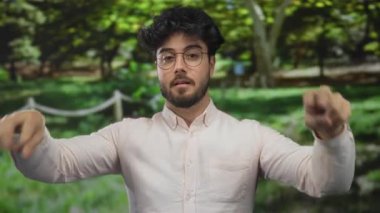 Sakallı, gözlüklü, beyaz gömlekli, güneşli yeşil park ortamında tırnak işareti yapan İspanyol bir genç..