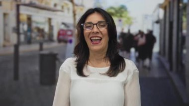 Gözlük takan ve gülümseyen dudaklı beyaz bluz giyen kadın, güneş ışığında kaldırım taşı caddesinde el çırpıyor..