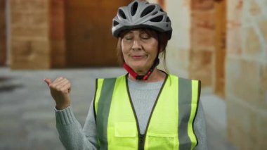 Senior hispanic woman wearing helmet and vest stands on city street, pointing to the side with a smile, blending safety and urban exploration.