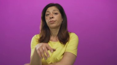 Stüdyoda eli çenesinde olan sarı gömlekli kadın mor duvara yaslanıyor; sakin yansıma.