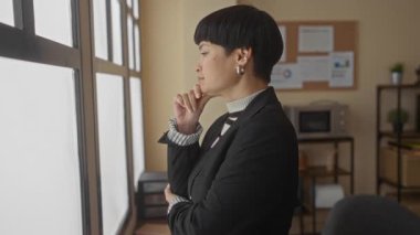 Kadın, resmi giyinmiş, parlak bir ofis penceresinin kenarında, arka planda dosyalar ve bir mikrodalga fırın ile modern bir iş yeri içerisini resmediyor..