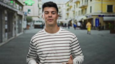 Çizgili tişörtlü genç adam canlı bir şehir sokak ortamında hoş bir jest yapıyor. Gün boyunca dışarıda dikilen kişinin kendine güveni ve neşesi artıyor..