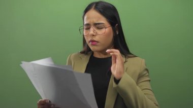 Kadın, yeşil arka planda dokümanları incelerken hayal kırıklığına uğramış görünüyor, boynunu ovalarken stres gösteriyor, gözlük takıyor ve bej ceket giyiyor..