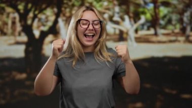 Parkta kutlama yapan neşeli bir kadın, sarı saçlı, gözlüklü, doğal ortamda neşe ve pozitifliği vurgulayan..