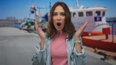 Deniz kenarındaki limanda sürpriz yapan genç bir kadın arka planda tekneler, canlı bir açık hava sahnesi çekiyor. Deniz ve turizm unsurları sergileniyor..